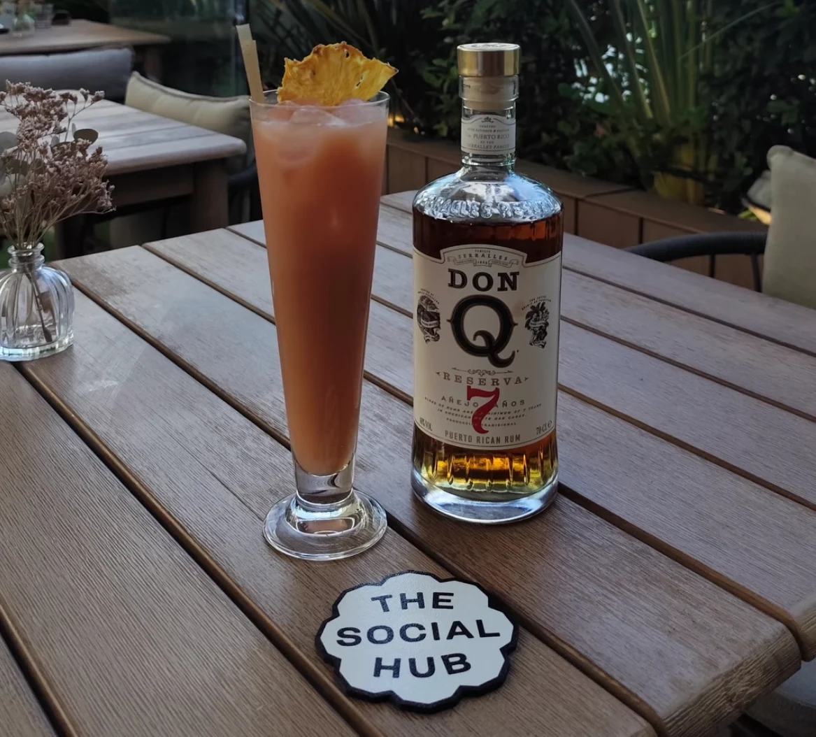 Posavasos The Social Hub junto a un cóctel alto y una botella sobre una mesa